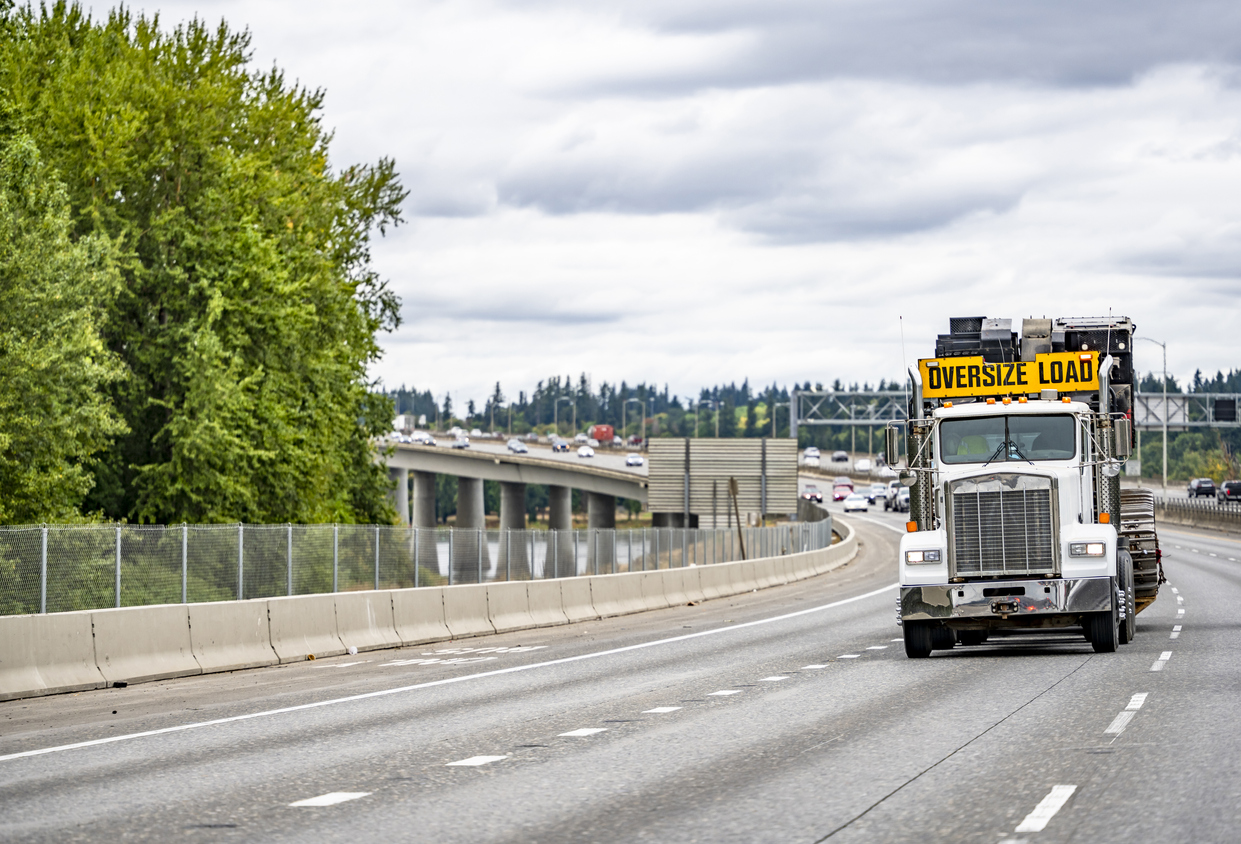 Powerful white big rig classic semi truck with oversize load sign on the roof transporting oversized cargo on step down semi trailer running on the winding overpass highway road across a River